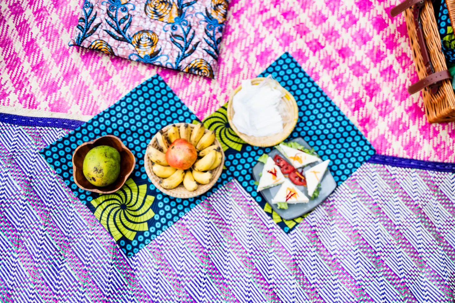 A picnic laid out on pink and blue woven mats with patterned cloths.